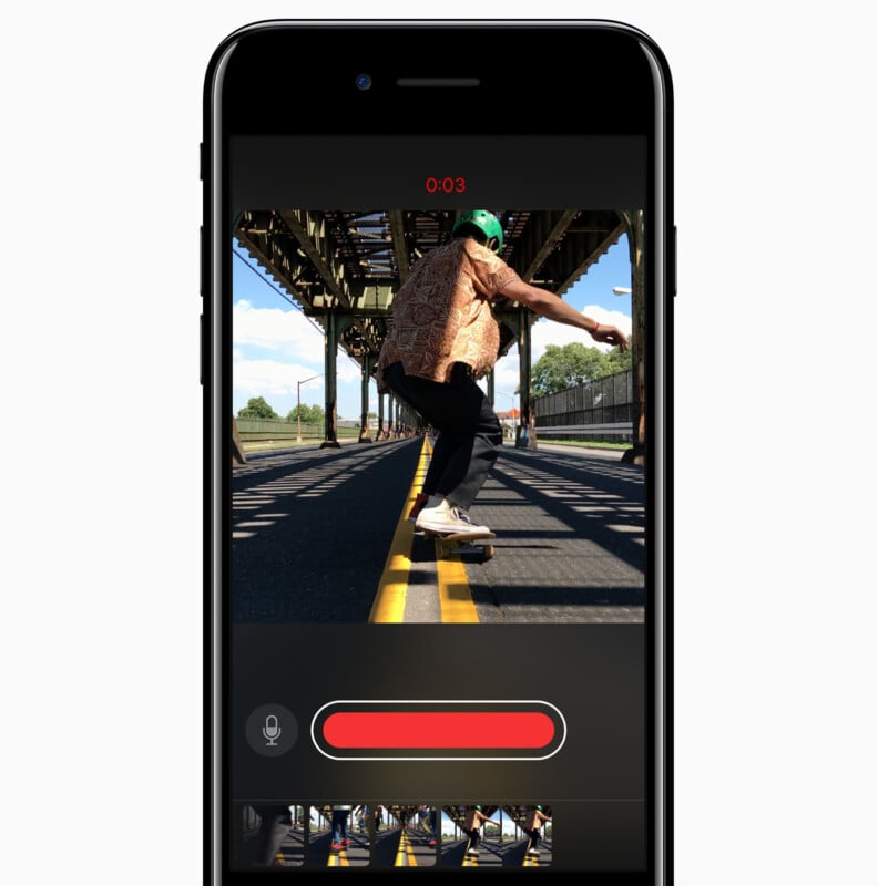 A smartphone screen displays a video recording of a person skateboarding under a metal bridge. The person is wearing a helmet and patterned shirt, and the street is lined with yellow lines.