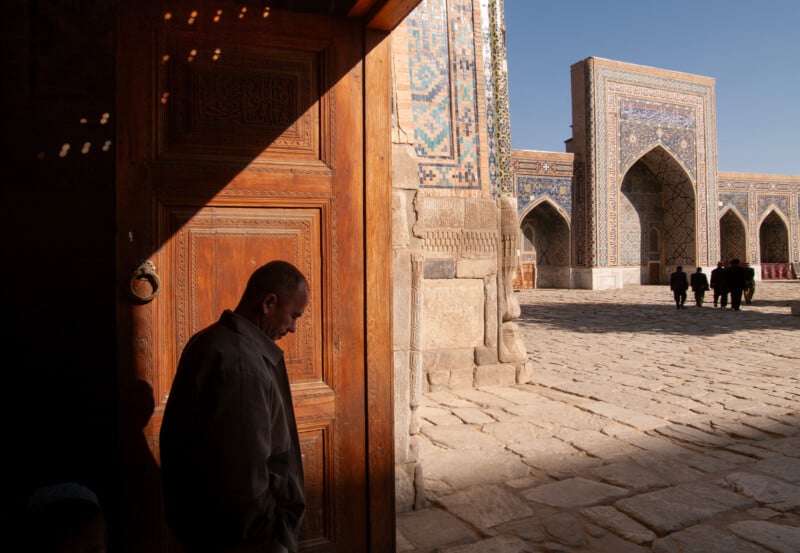 A man stands in shadow by an ornate wooden door, with sunlight illuminating a courtyard of an intricately tiled building; several people walk in the distance under blue sky.