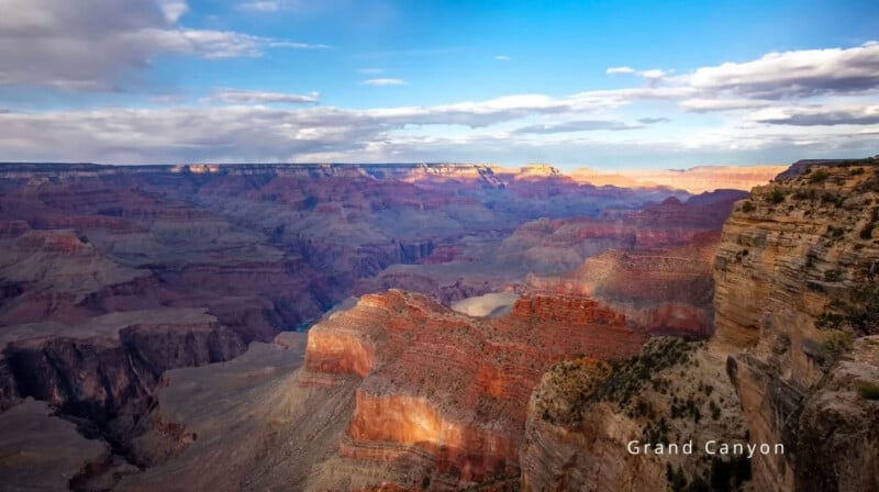 Bajo un cielo nublado, una vista panorámica del Gran Cañón muestra coloridas formaciones rocosas en capas, cañones profundos y acantilados escarpados. El texto en la esquina inferior derecha dice "Gran Cañón.