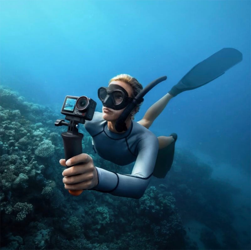 A person wearing snorkeling gear swims underwater near a coral reef, holding an action camera on a handle to capture footage in the clear blue ocean.