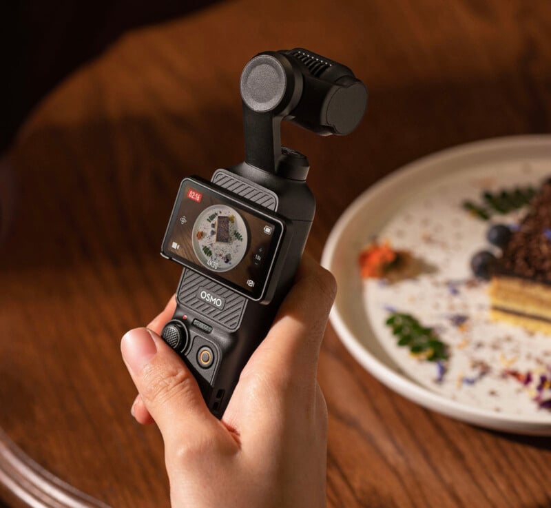 A hand holds a DJI Osmo Pocket 3 camera, pointed at a dessert on a white plate with blueberries and garnish, placed on a wooden table. The camera screen displays the dessert being recorded.