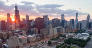 Aerial view of a city skyline at sunset, with sunlight reflecting off tall skyscrapers and clouds in the sky. Streets and green spaces are visible in the foreground.