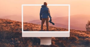 A person holding a camera stands on a grassy hill at sunset, with an oversized computer monitor frame highlighting them and the mountain landscape in the background.