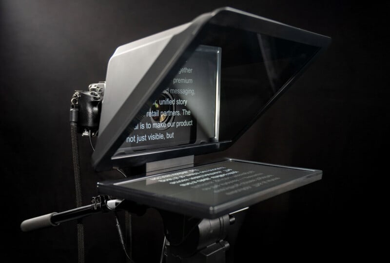 A close-up of a teleprompter setup, showing a camera behind angled reflective glass displaying scrolling text, used for presenters to read scripts while looking into the camera.