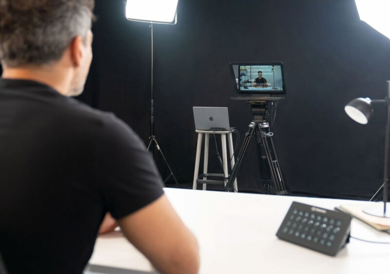 A person sits at a desk in a video production studio with professional lights and a camera on a tripod. A laptop is on a stool, and the camera screen shows the person’s image being recorded.