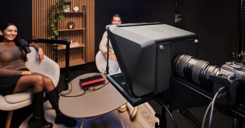 Two people sit in a modern recording studio, speaking into microphones. A camera with a teleprompter setup is positioned in the foreground, capturing the conversation. A shelf with books and plants is in the background.