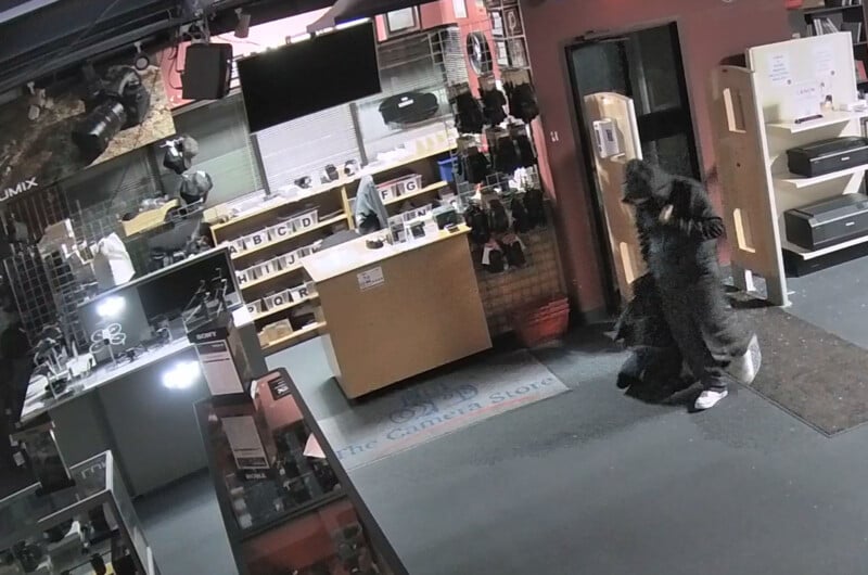 A person wearing a black costume or coat, possibly with a mask, walks through a store filled with camera equipment and accessories. The store is lit and has displays and shelves along the walls.