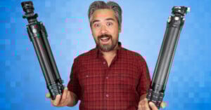 A man in a red plaid shirt stands in front of a blue pixelated background, smiling and holding a tripod in each hand.
