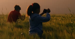 Two people sit in a field of tall grass at sunset; the person in the foreground wears a blue jacket and takes a photo, while the person in the background, in red, looks down at something in their hands.