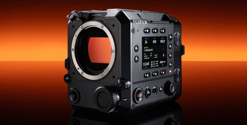 A black Fujifilm digital cinema camera with a large lens mount and multiple control buttons is displayed on a glossy surface, with an orange gradient background. The side screen shows camera settings and status information.