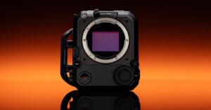 A compact, black professional camera body with exposed sensor, various control dials, and a large side grip, displayed against an orange gradient background.