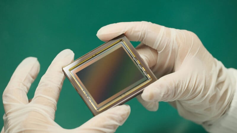 Gloved hands hold a rectangular camera sensor or electronic component with a reflective surface against a green background.