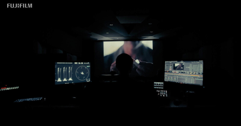 Una persona está sentada en una habitación oscura, frente a varios monitores de computadora, editando videoclips, con una gran pantalla borrosa de fondo. "fujifilm" Texto en la esquina superior izquierda.