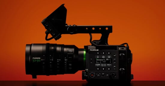 A professional cinema camera with a large Fujinon lens is displayed in profile against a vibrant orange background. The camera features various control buttons and a top handle.