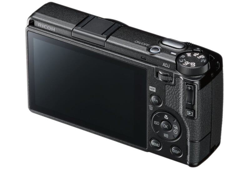 A black Ricoh digital camera is shown from the rear, displaying its large LCD screen, control buttons, and mode dial. The camera's textured grip and compact design are visible.