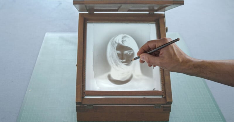 A hand holding a pencil traces over a photographic negative of a woman's face, which is placed inside a wooden box with a glass lid. The box sits on a green cutting mat.