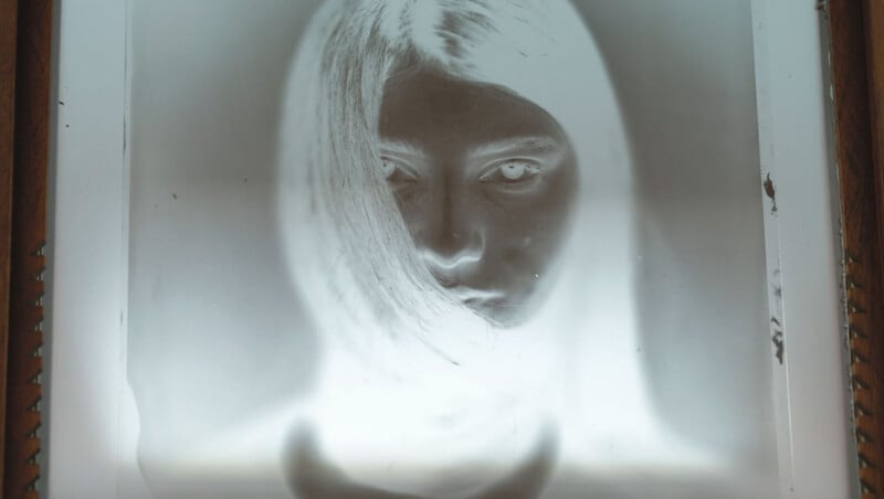 A black-and-white photographic negative of a person with long hair partially covering their face, staring directly at the camera. The image has high contrast and visible film edges.