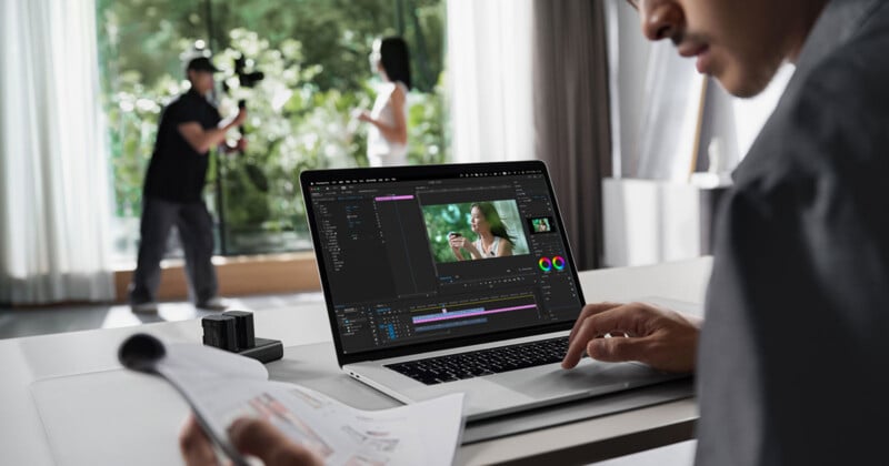 Un hombre edita un vídeo en una computadora portátil en un escritorio, mientras que en el fondo un camarógrafo filma a una mujer parada cerca de una gran ventana en una habitación luminosa.