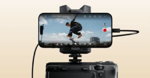 A smartphone mounted on a camera records a skateboarder mid-jump against a blue sky with clouds and distant buildings, with camera interface icons visible on the phone's screen.