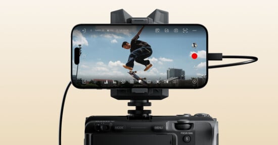 A smartphone mounted on a camera records a skateboarder mid-jump against a blue sky with clouds and distant buildings, with camera interface icons visible on the phone's screen.