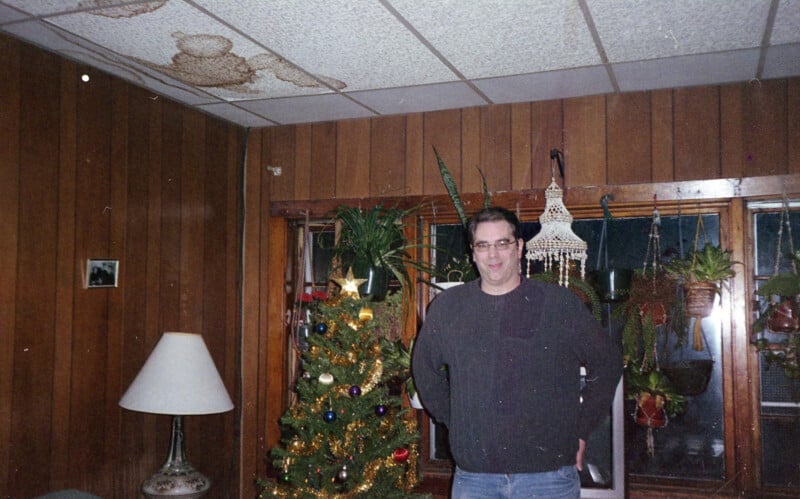 A man wearing glasses and a dark sweater stands in a wood-paneled room beside a decorated Christmas tree, with hanging plants and a lamp in the background. The ceiling shows noticeable water stains.