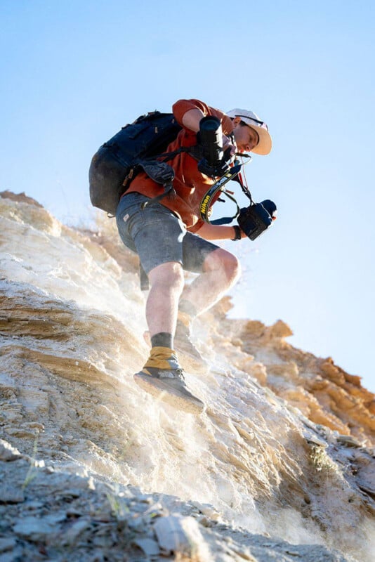 A person wearing a hat and backpack carefully descends a steep, dusty rocky slope while holding a camera in each hand under a clear blue sky.