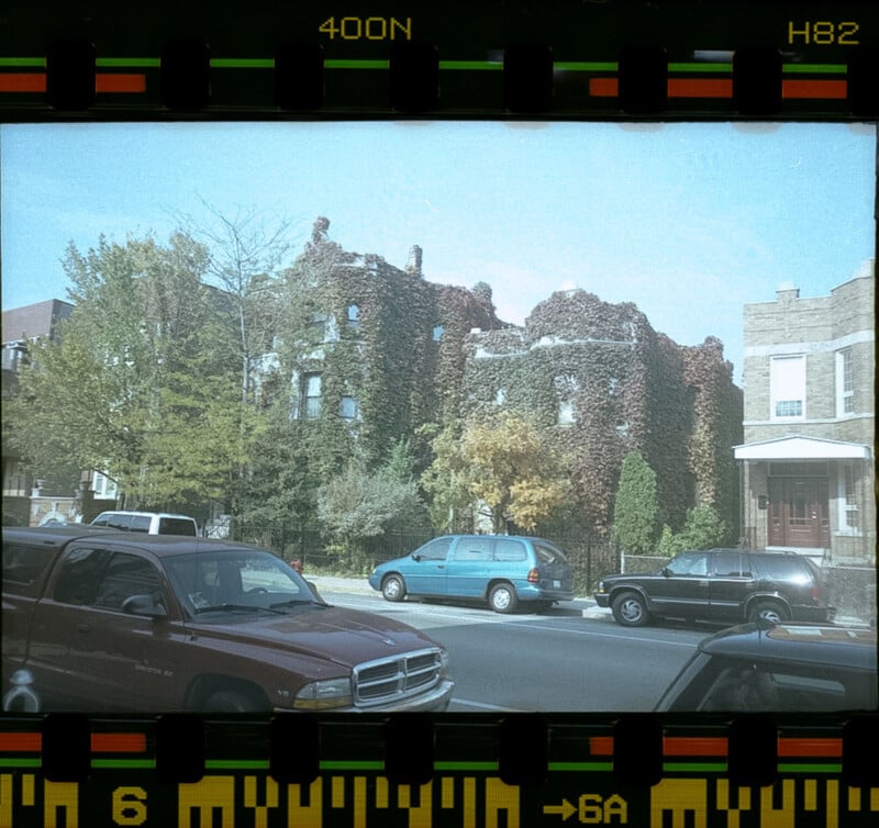 A residential street with parked cars in front of a house covered in dense ivy, surrounded by trees. The photo has a film strip border and a vintage, faded appearance.