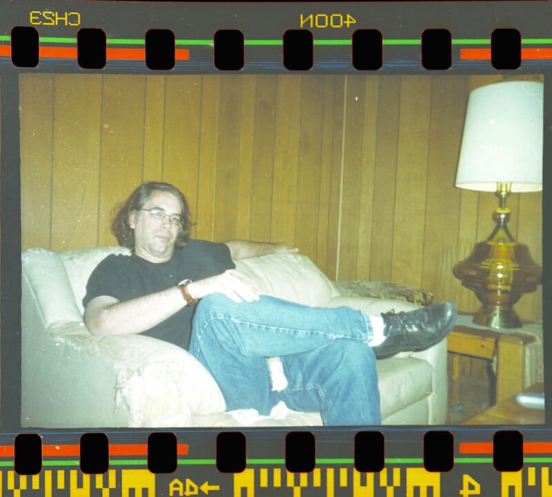 A man with glasses and long hair sits casually on a beige couch in a wood-paneled room. He is wearing a black shirt and jeans, with one leg crossed over the other. A lamp and side table are nearby. The image resembles a film negative.