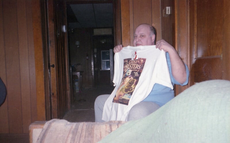 A man sitting in a wood-paneled room holds up a white T-shirt featuring a "Ghostbusters" graphic. The room has dim lighting, a hallway in the background, and part of a green blanket is visible in the foreground.