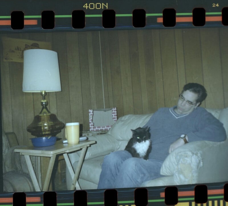 A man sits on a beige couch in a wood-paneled room, looking down at a black and white cat on his lap. A lamp and a yellow cup are on a small table beside him. The image has a film strip border.