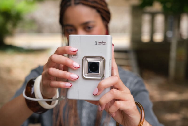 Un hombre con coletas sostiene una cámara instantánea Instax Mini blanca cerca de su cara, apuntando con la lente al espectador. Tenían las uñas pintadas de rosa pálido y llevaban gruesas pulseras. El fondo es borroso y al aire libre.