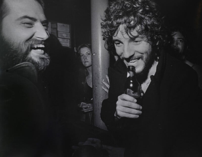 Dos hombres ríen juntos en un bar con poca luz; uno de ellos se lleva una botella de cerveza a los labios. Al fondo había una mujer y un hombre, todos parecían relajados e inmersos en la animada atmósfera.