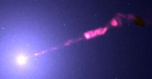A bright celestial object emits a glowing pink jet of light extending diagonally through space, surrounded by faint stars against a dark background.