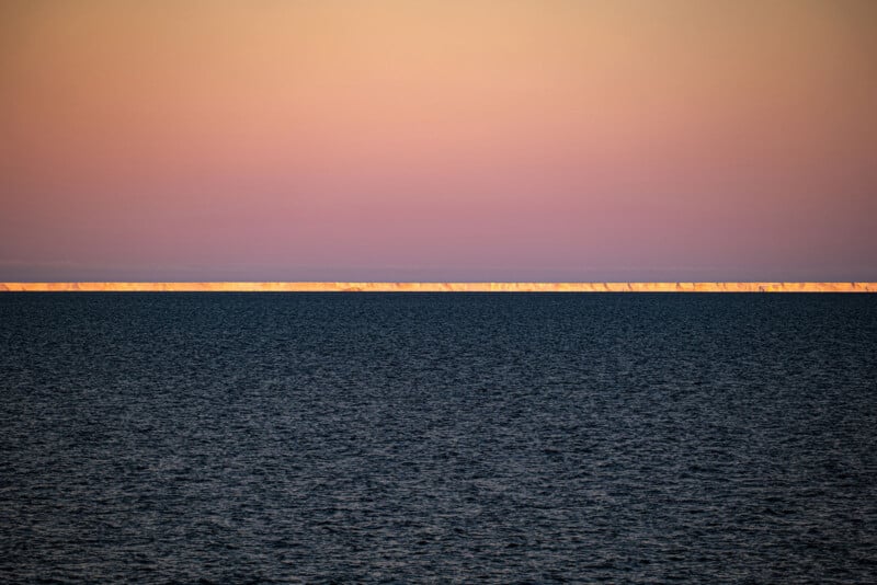A distant, sunlit ice shelf glows on the horizon beneath a pastel pink and orange sky over a dark, calm ocean.