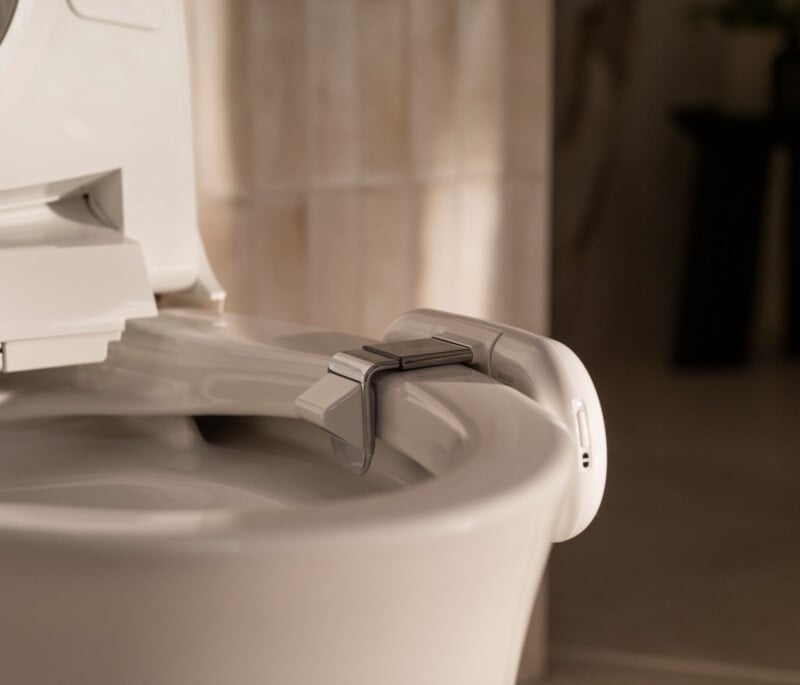 A close-up of a white toilet with a bidet attachment installed on the rim, showing control features on the side in a softly lit bathroom.