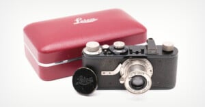 A vintage Leica camera with a lens cap beside it sits in front of a red Leica-branded case on a white surface.
