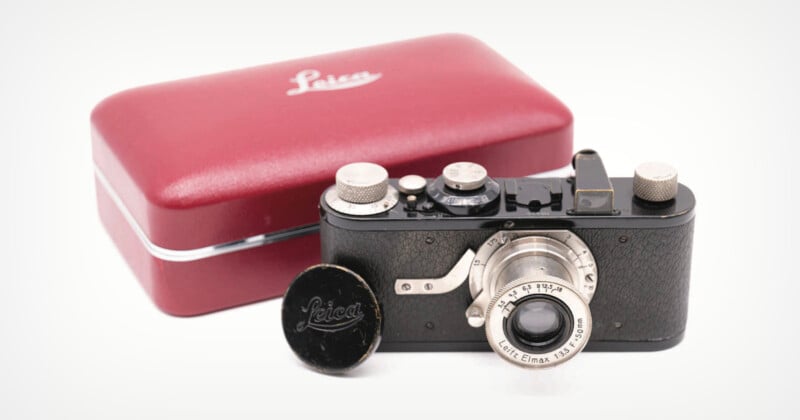 A vintage Leica camera with a lens cap beside it sits in front of a red Leica-branded case on a white surface.