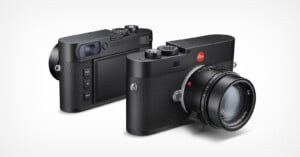 Two black Leica digital cameras are shown, one facing forward displaying the lens and the other turned to show the back with its screen and control buttons. Both cameras have a textured grip and classic, minimalist design.
