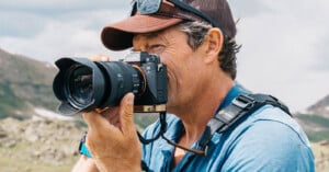 A man wearing a cap and sunglasses on his head holds a camera up to his face, taking a photo outdoors with mountains and a cloudy sky in the background.