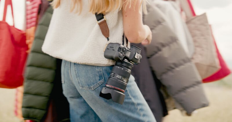 A person with blonde hair carries several bags and a camera hanging from a strap over their shoulder, wearing blue jeans and a white sleeveless top. The background is softly blurred.