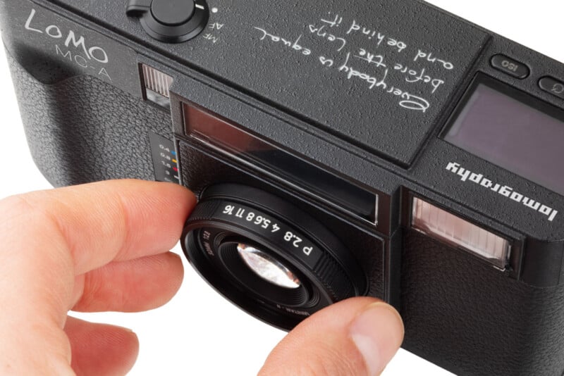 A close-up of a person adjusting the lens on a black LOMO LC-A camera, with handwritten text in white ink visible on the camera body near the viewfinder.