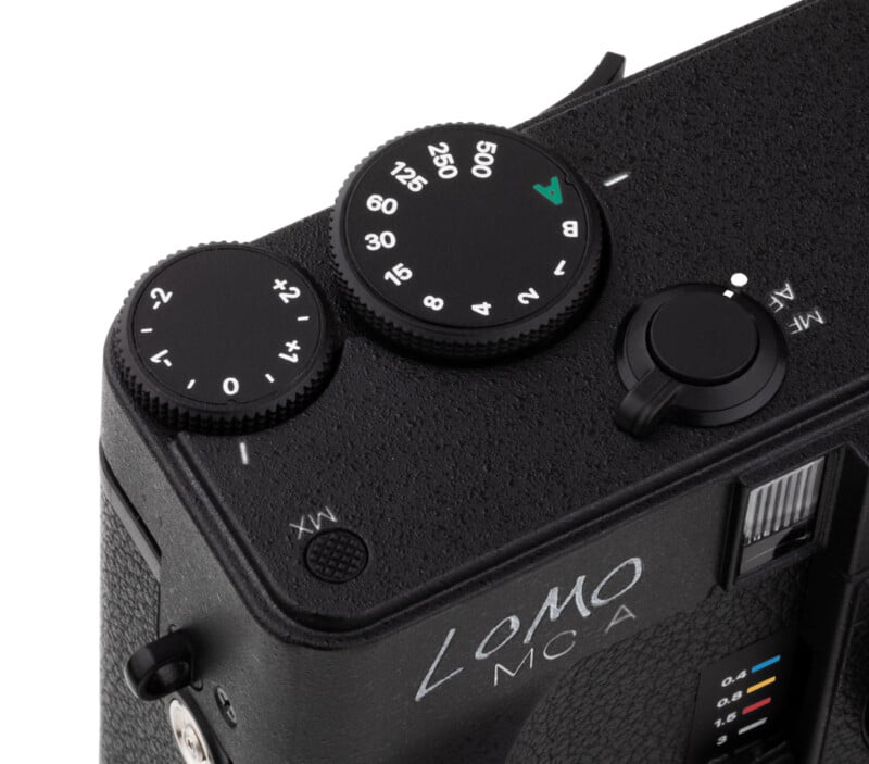 Close-up of the top corner of a black LOMO camera, showing textured dials for aperture and shutter speed, with visible numbers and a small silver button beside the "LOMO MCA" branding.