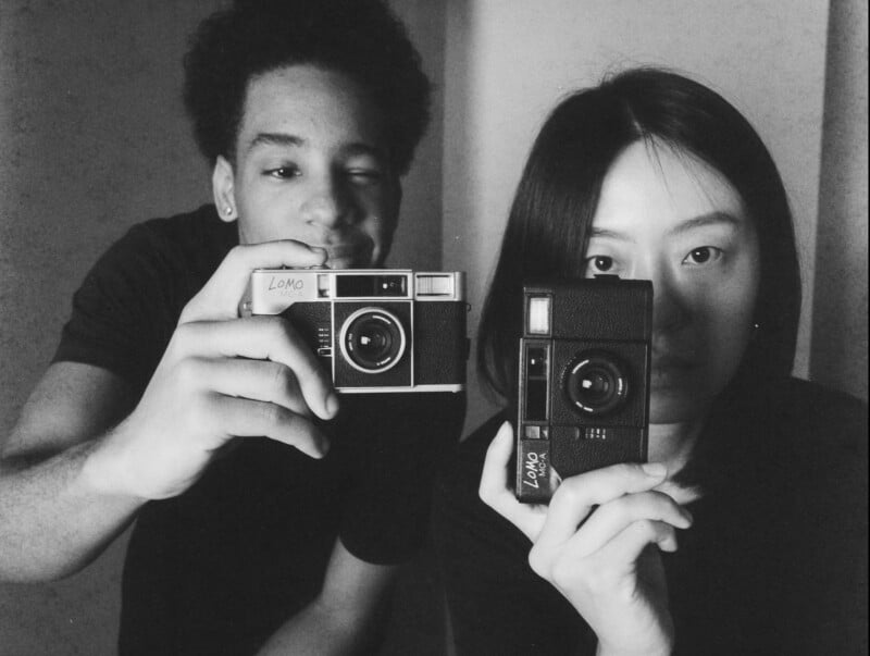 Two people stand close together, each holding up a vintage film camera toward the mirror. One person winks while the other looks directly into the lens. The photo is in black and white.