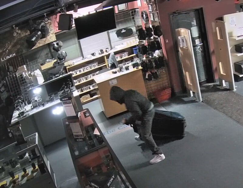 A person in a hooded jacket and mask holds a large black bag inside a camera store at night, appearing to steal items. Glass display cases and camera equipment are visible around them. The store looks disorganized.