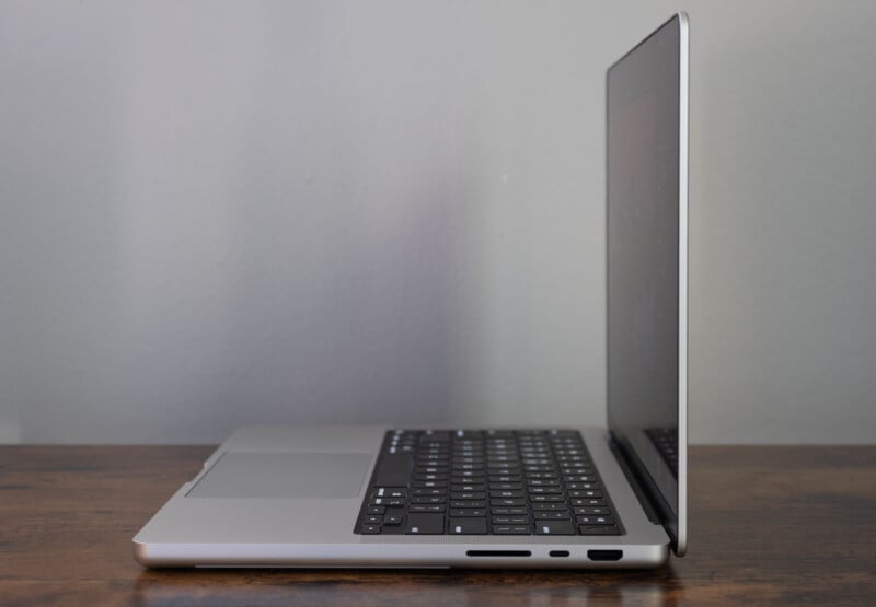 A silver laptop sits open on a wooden surface, viewed from the side. The screen is partially open, and various ports are visible along the side of the laptop. The background is a plain light gray wall.