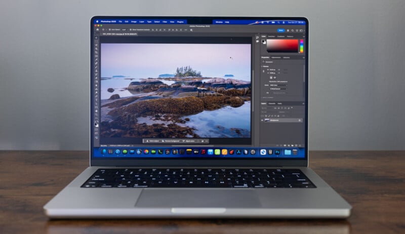 A laptop on a wooden table displays a photo editing software with an image of a rocky shoreline and small island, showing editing tools and adjustment panels on the screen.