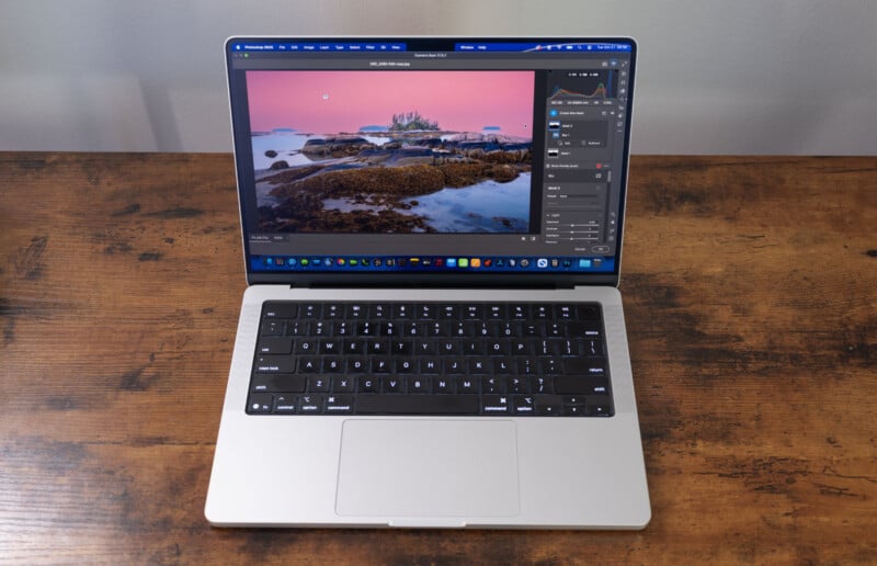 A silver laptop on a wooden table displays photo editing software with an image of rocks and water at sunset on the screen. The keyboard and touchpad are visible.