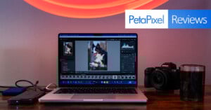 A laptop displaying a photo-editing program with a dog image on screen sits on a desk next to a camera, glass, and hard drive. The "PetaPixel Reviews" logo is in the top right corner against a lit wall.
