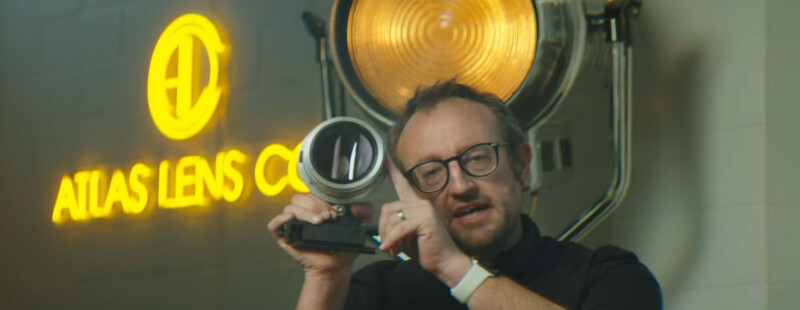 Un hombre con gafas se para frente a grandes luces de estudio y letreros de neón, sosteniendo la lente de la cámara y haciendo gestos "lente atlas corp.