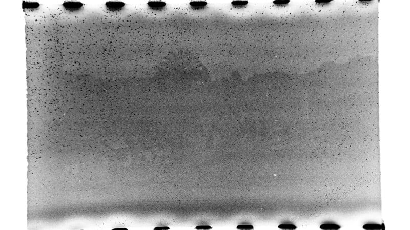 A black and white film negative with sprocket holes along the top and bottom edges, featuring heavy grain, speckles, and faint, blurry shapes, making the image content unclear.